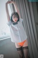 A woman in a white shirt and orange panties posing by a window.